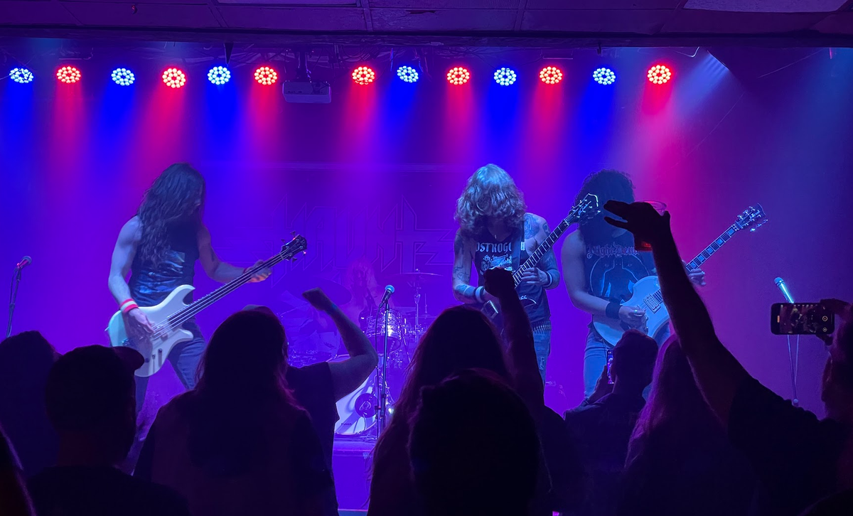 Three guitarists on stage, with red and blue lights above them, and a silhouette of an audience in the the foreground.